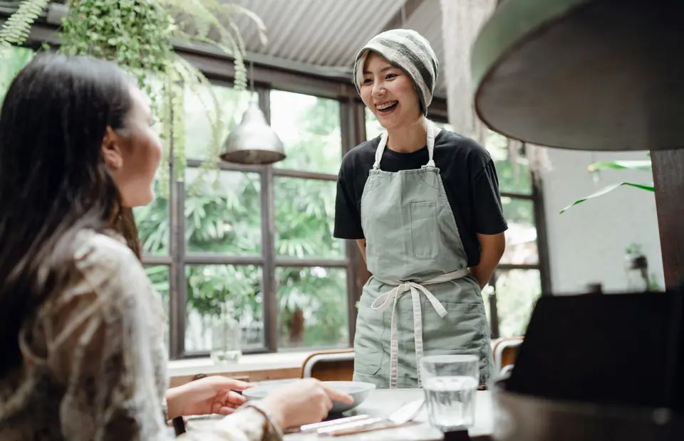 Lachende Servicekraft mit Schürze steht an einem Tisch im Café und spricht mit einem Gast